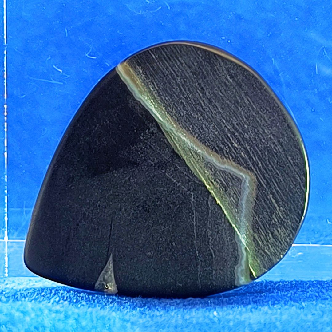 Plettro in pietra di onice nera con striature bianche trasparenti e adesivo con micro granuli di quarzo per ottimizzare il grip e la presa sul codolo del plettro, fotografato con sfondo azzurro.
						