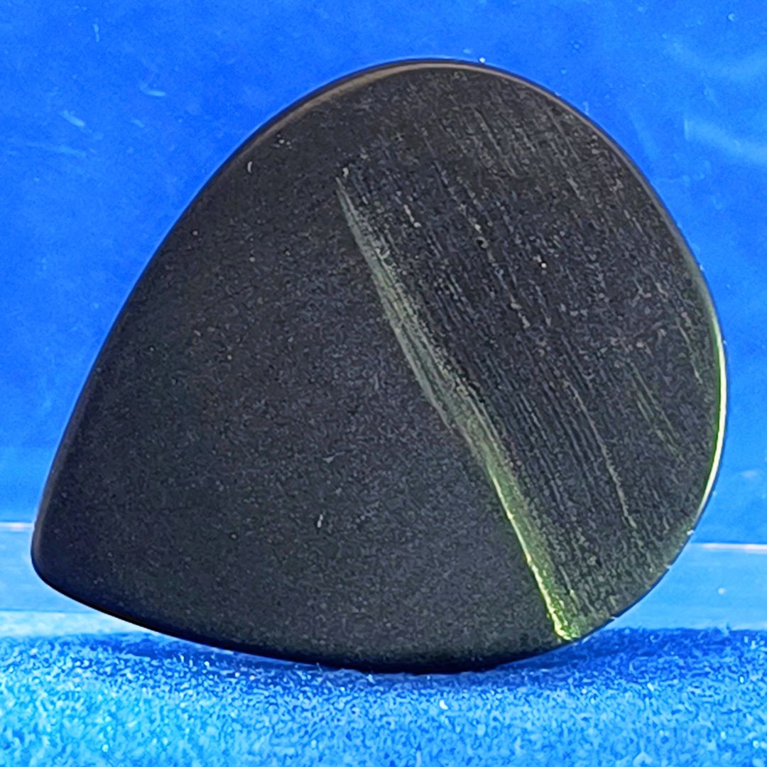 Plettro in pietra di onice nera con striature bianche trasparenti e adesivo con micro granuli di quarzo per ottimizzare il grip e la presa sul codolo del plettro, fotografato con sfondo azzurro.
						