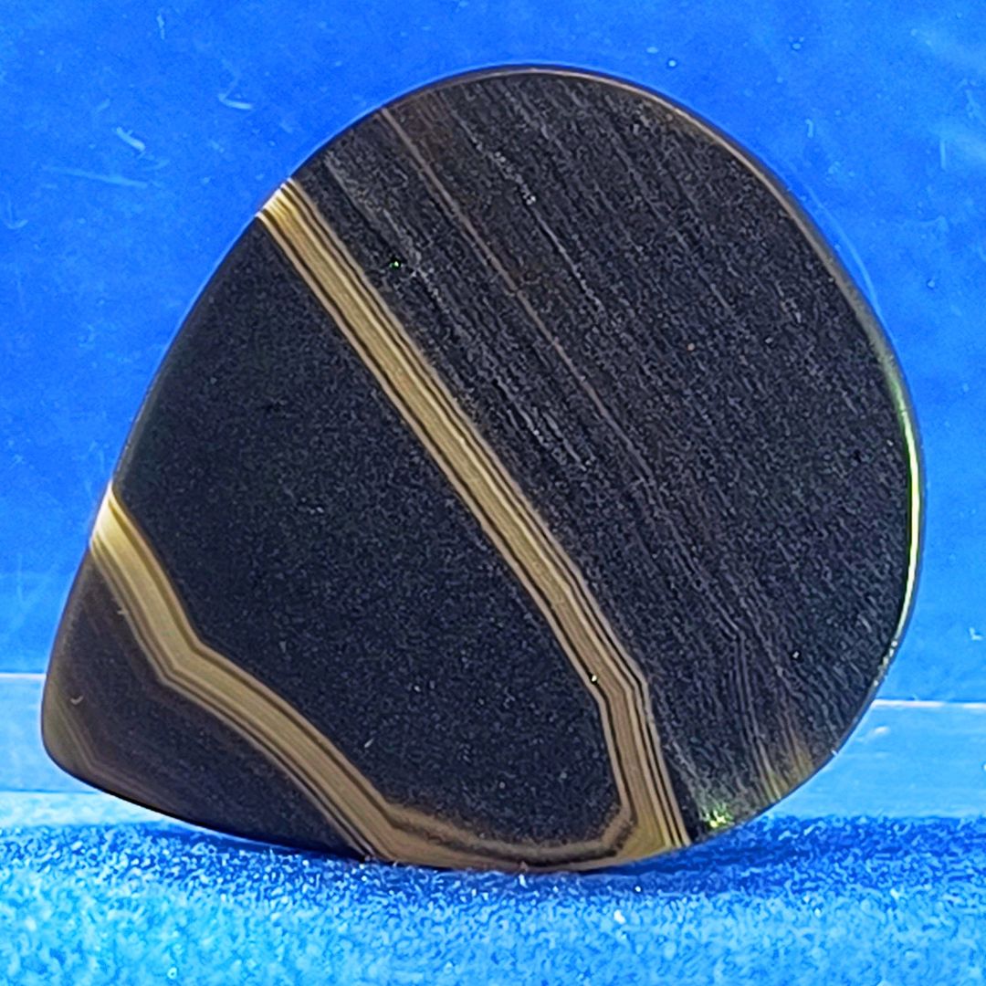 Plettro in pietra di onice nera con striature bianche trasparenti e adesivo con micro granuli di quarzo per ottimizzare il grip e la presa sul codolo del plettro, fotografato con sfondo azzurro.