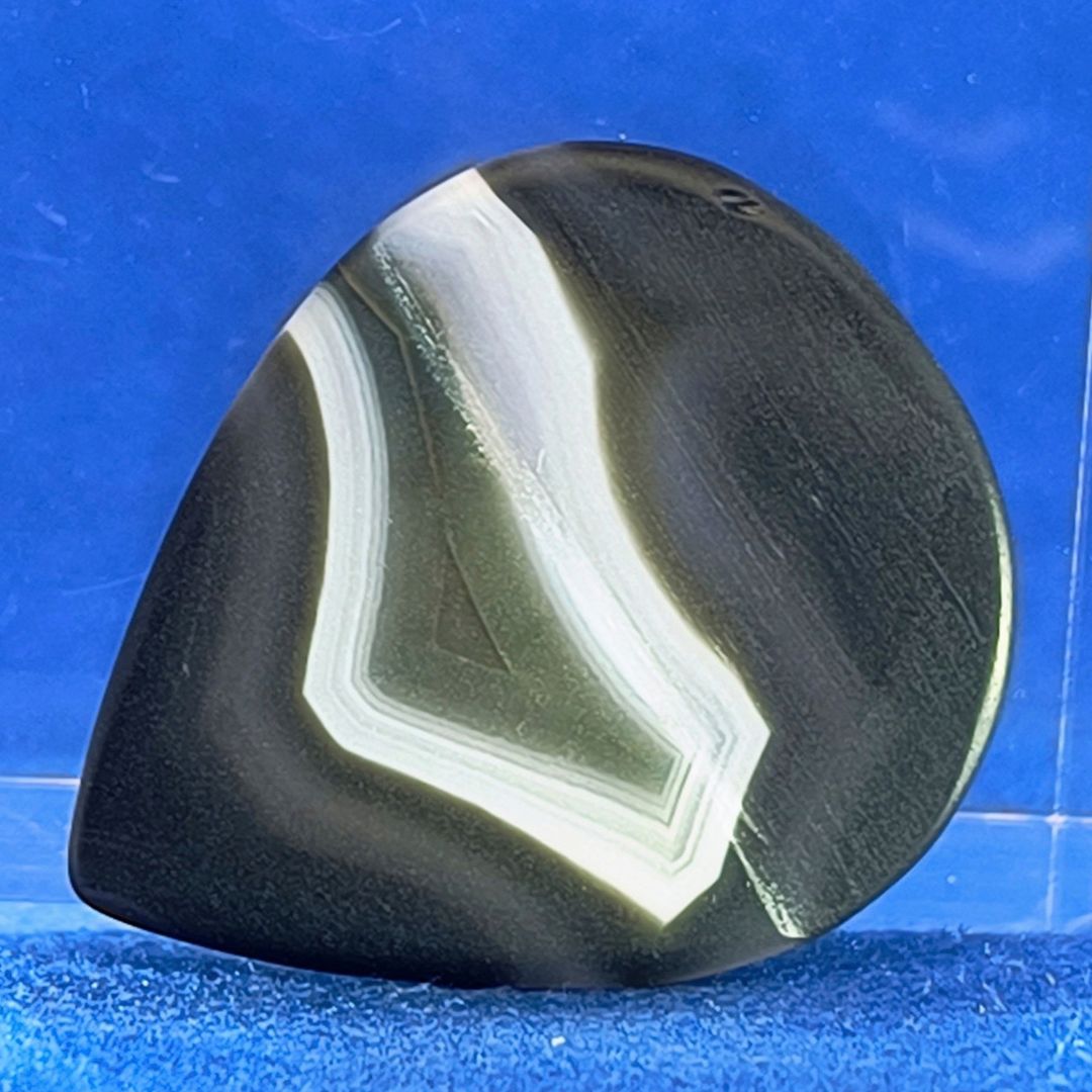 Plettro in pietra di onice nera con striature bianche trasparenti e adesivo con micro granuli di quarzo per ottimizzare il grip e la presa sul codolo del plettro, fotografato con sfondo azzurro.