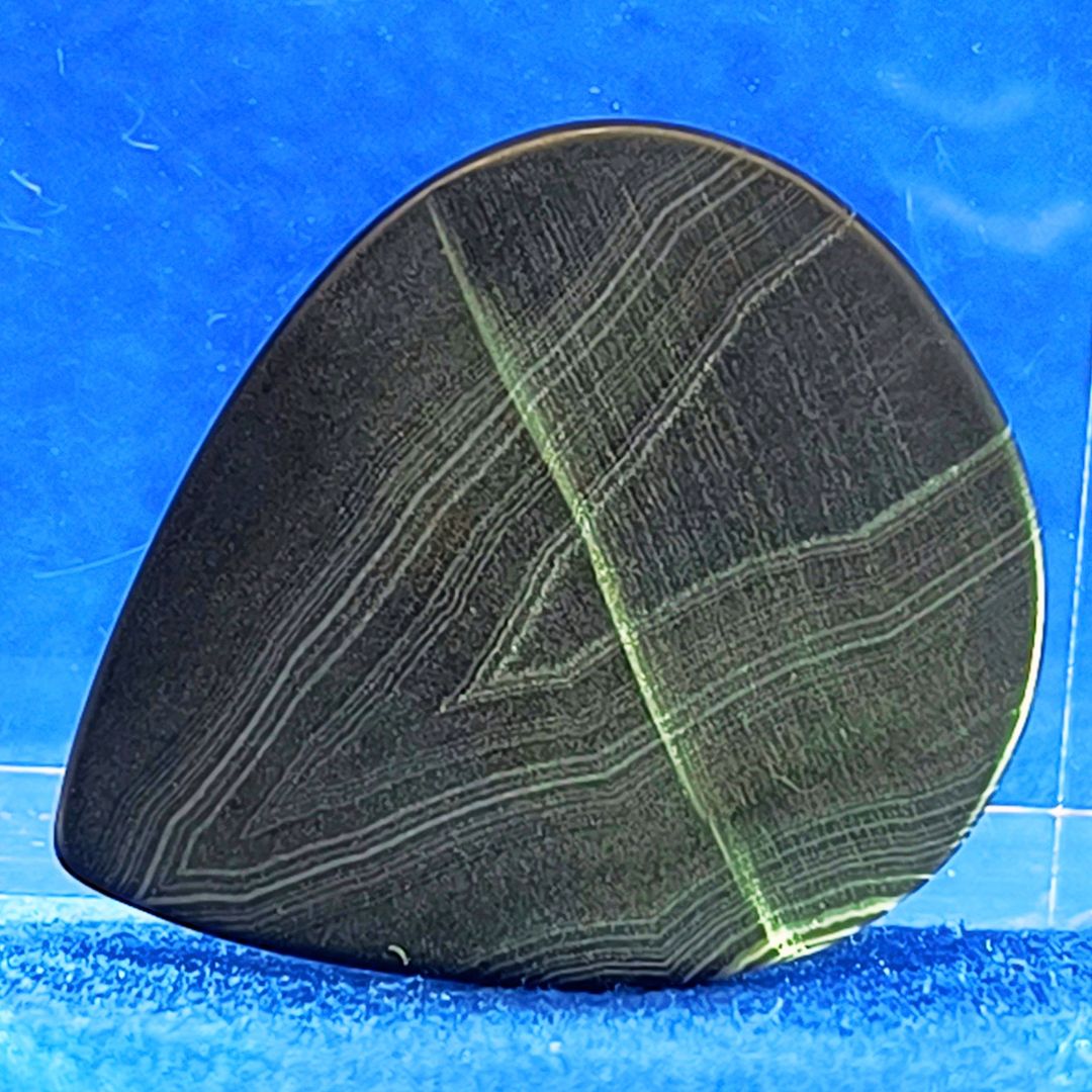 Plettro in pietra di onice nera con striature bianche trasparenti e adesivo con micro granuli di quarzo per ottimizzare il grip e la presa sul codolo del plettro, fotografato con sfondo azzurro.