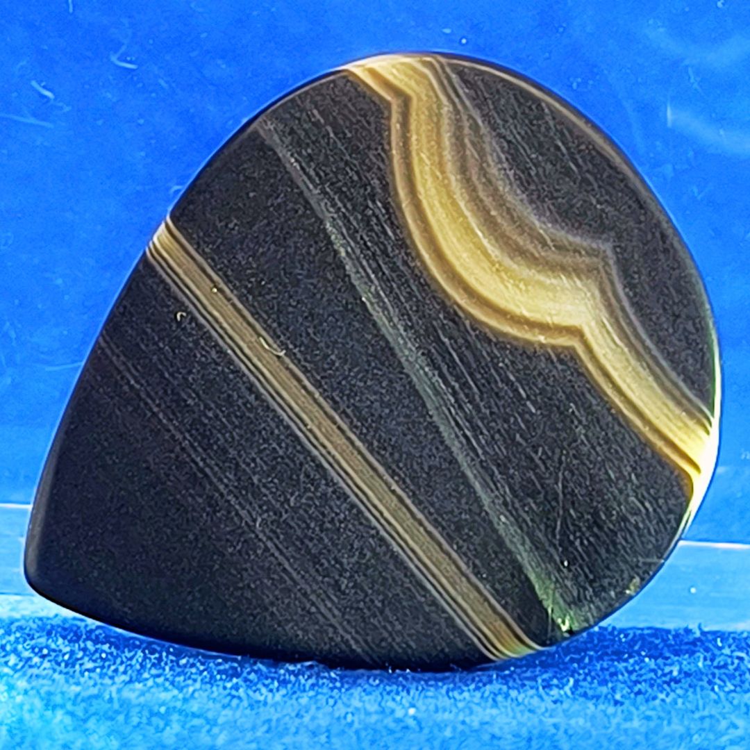 Plettro in pietra di onice nera con striature bianche trasparenti e adesivo con micro granuli di quarzo per ottimizzare il grip e la presa sul codolo del plettro, fotografato con sfondo azzurro.