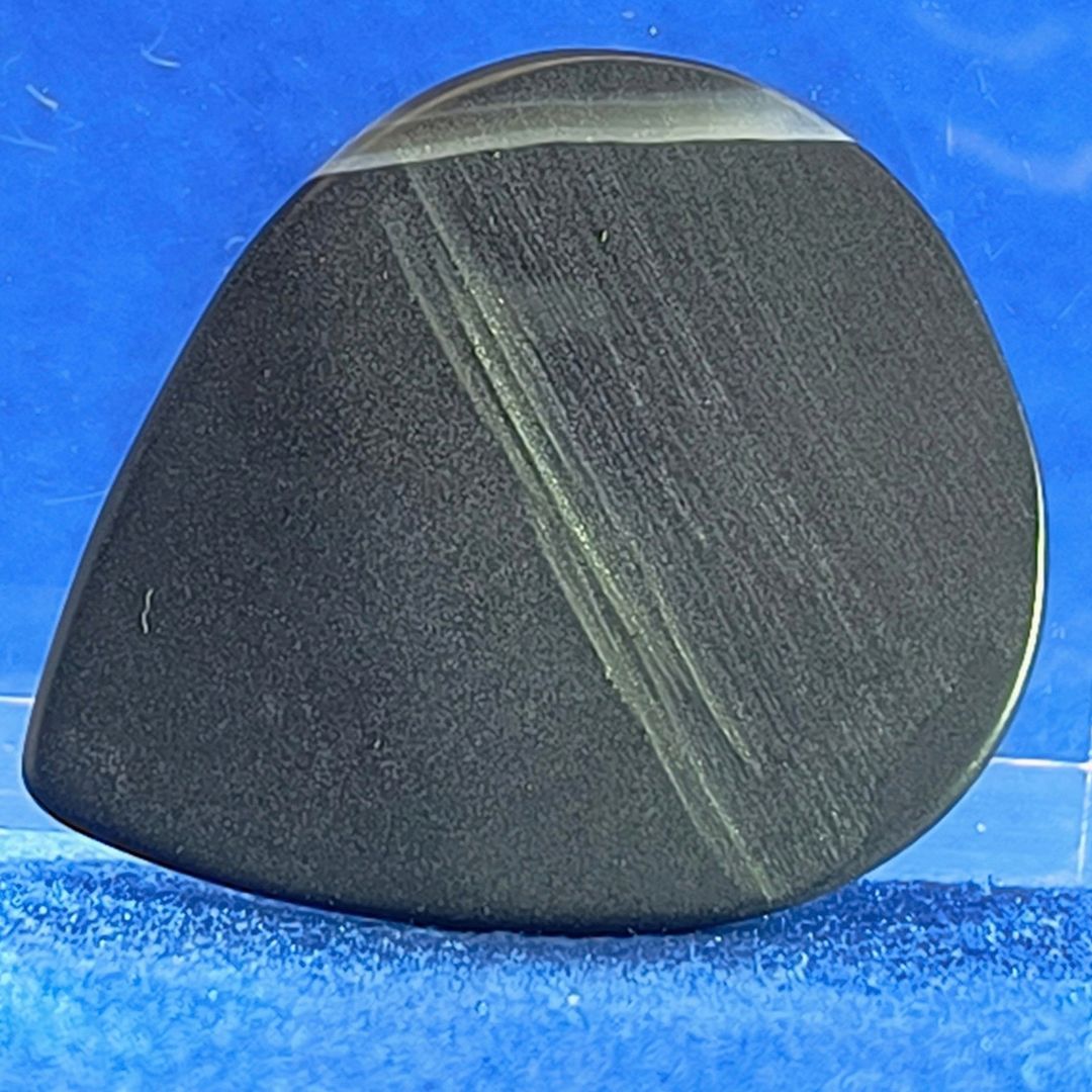 Plettro in pietra di onice nera con striature bianche trasparenti e adesivo con micro granuli di quarzo per ottimizzare il grip e la presa sul codolo del plettro, fotografato con sfondo azzurro.
						
						