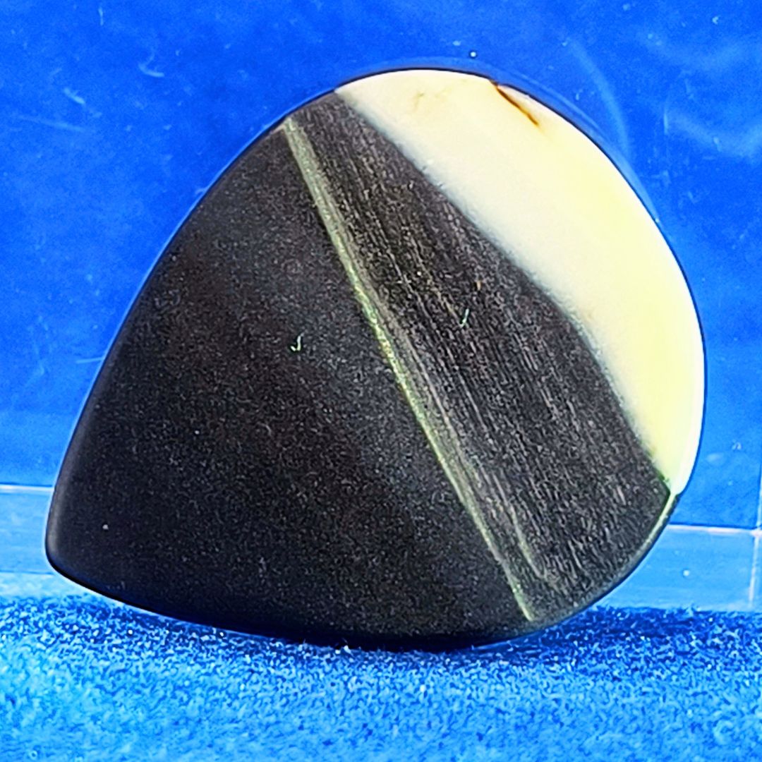 Plettro in pietra di onice nera con striature bianche trasparenti e adesivo con micro granuli di quarzo per ottimizzare il grip e la presa sul codolo del plettro, fotografato con sfondo azzurro.