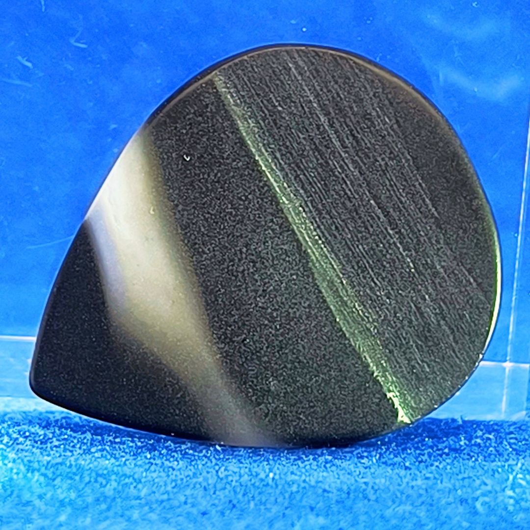 Plettro in pietra di onice nera con striature bianche trasparenti e adesivo con micro granuli di quarzo per ottimizzare il grip e la presa sul codolo del plettro, fotografato con sfondo azzurro.