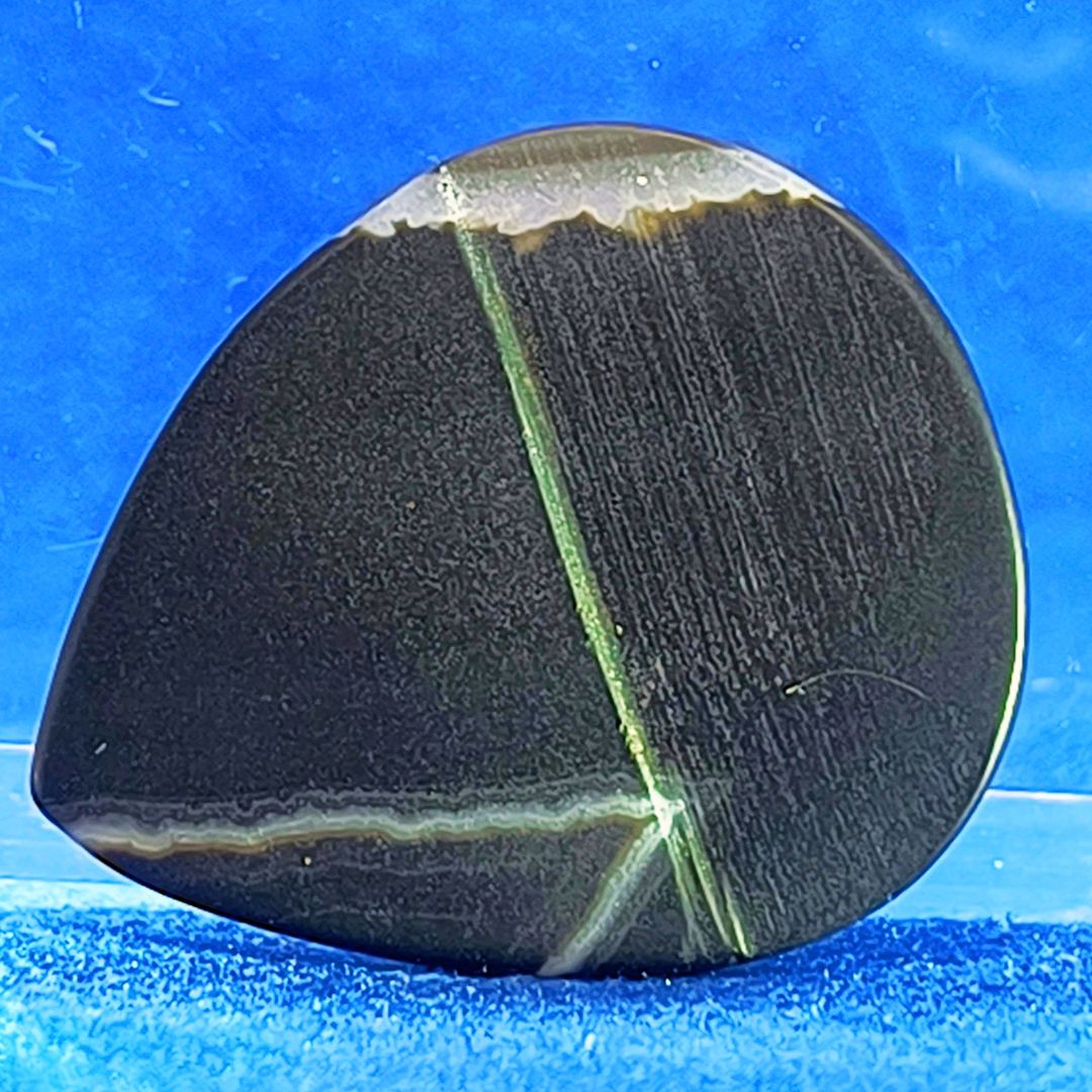 Plettro in pietra di onice nera con striature bianche trasparenti e adesivo con micro granuli di quarzo per ottimizzare il grip e la presa sul codolo del plettro, fotografato con sfondo azzurro.