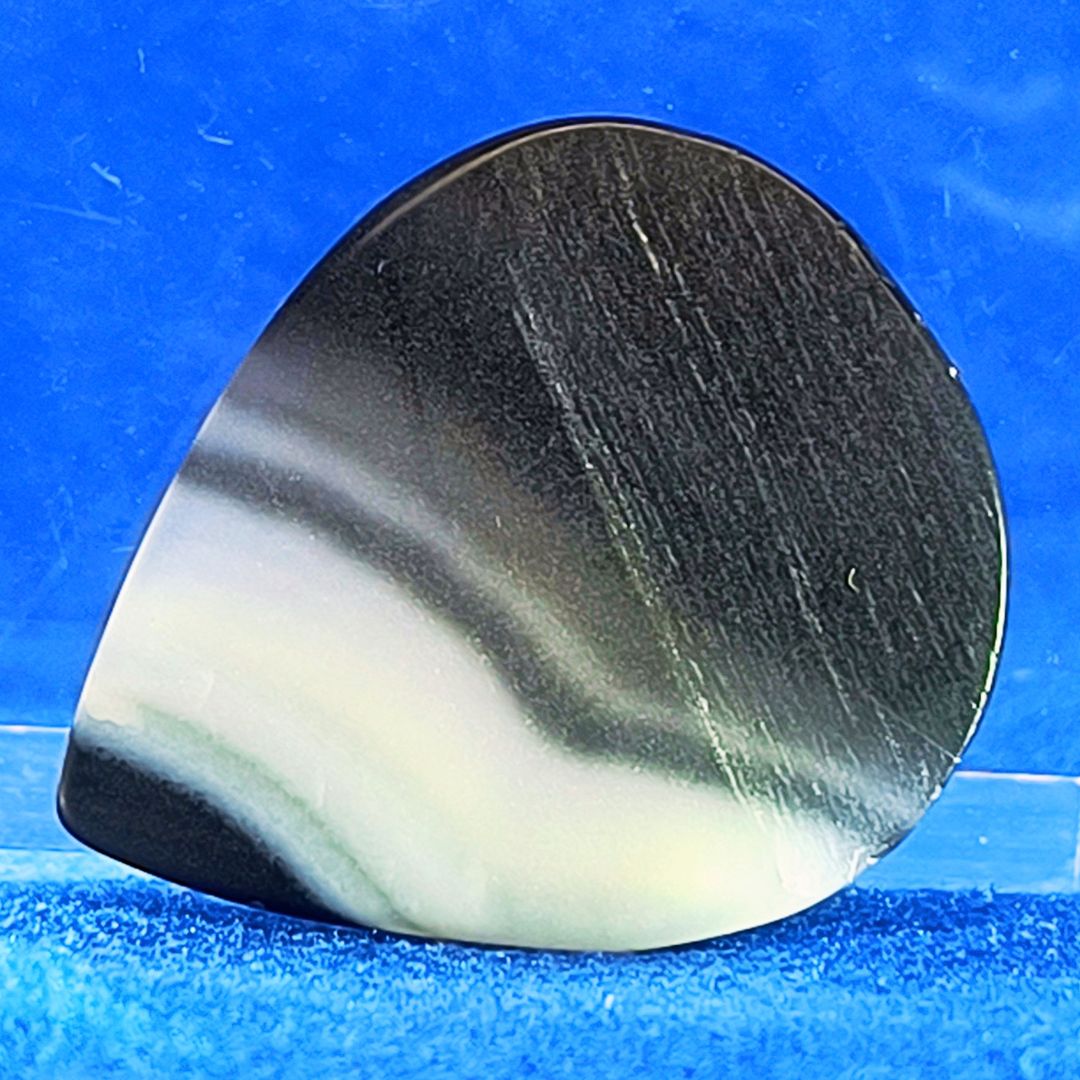 Plettro in pietra di onice nera con striature bianche trasparenti e adesivo con micro granuli di quarzo per ottimizzare il grip e la presa sul codolo del plettro, fotografato con sfondo azzurro.