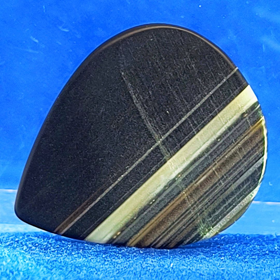 Plettro in pietra di onice nera con striature bianche trasparenti e adesivo con micro granuli di quarzo per ottimizzare il grip e la presa sul codolo del plettro, fotografato con sfondo azzurro.