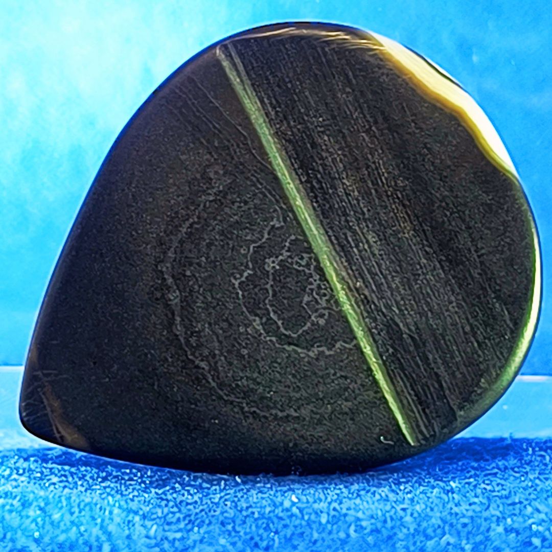 Plettro in pietra di onice nera con striature bianche trasparenti e adesivo con micro granuli di quarzo per ottimizzare il grip e la presa sul codolo del plettro, fotografato con sfondo azzurro.
