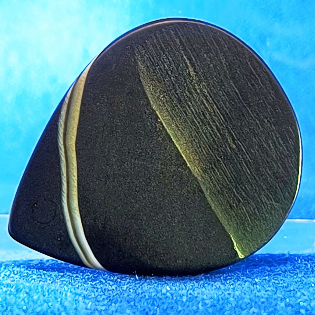 Plettro in pietra di onice nera con striature bianche trasparenti e adesivo con micro granuli di quarzo per ottimizzare il grip e la presa sul codolo del plettro, fotografato con sfondo azzurro.
						