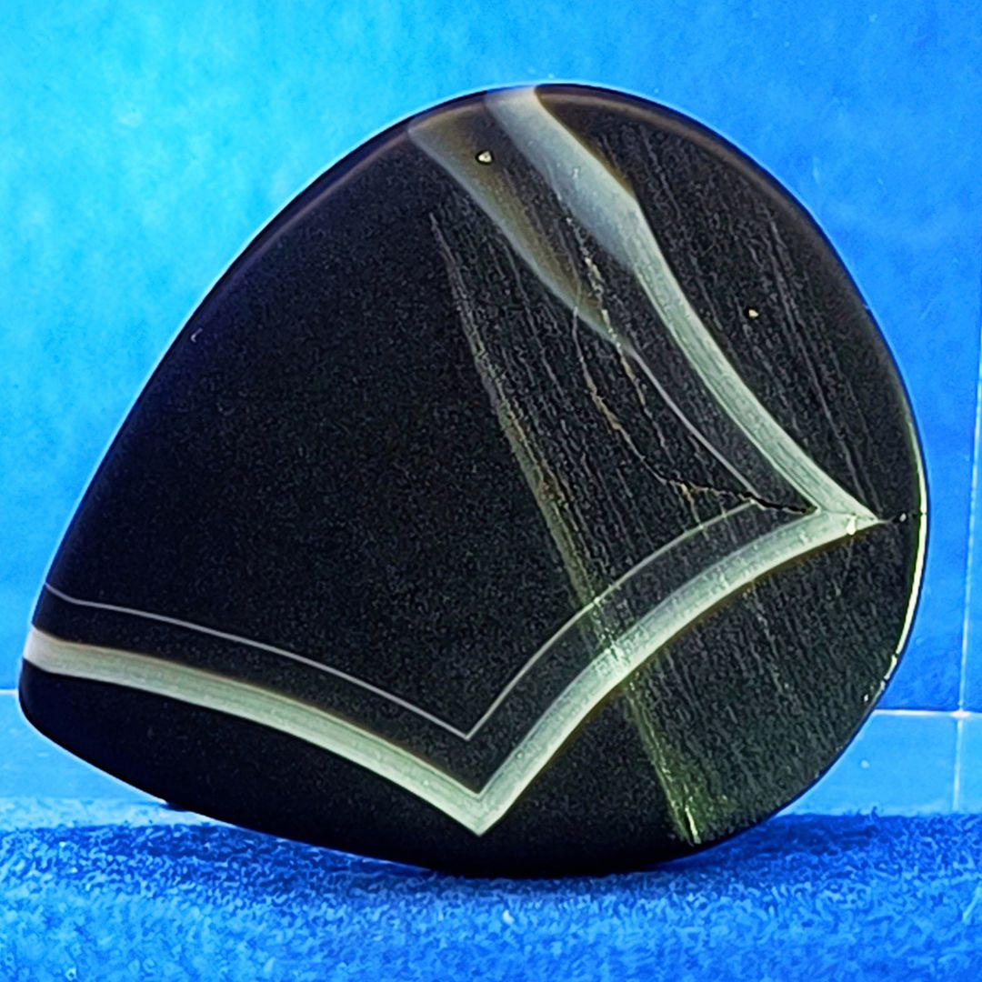Plettro in pietra di onice nera con striature bianche trasparenti e adesivo con micro granuli di quarzo per ottimizzare il grip e la presa sul codolo del plettro, fotografato con sfondo azzurro.
						