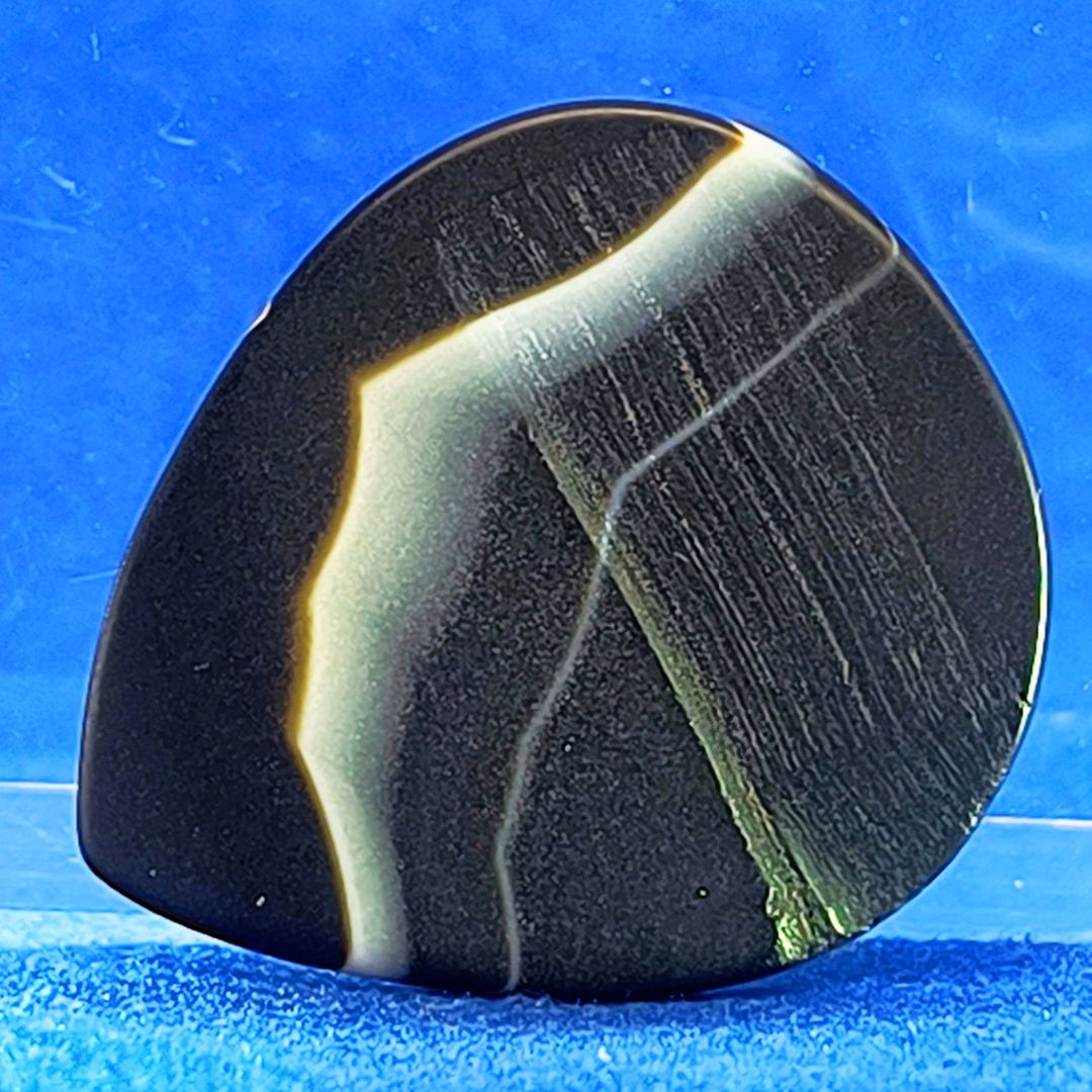 Plettro in pietra di onice nera con striature bianche trasparenti e adesivo con micro granuli di quarzo per ottimizzare il grip e la presa sul codolo del plettro, fotografato con sfondo azzurro.